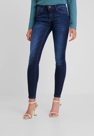Jeans ajustados de denim azul oscuro con un leve desgastado, diseño de cinco bolsillos y cintura media. Combinados con sandalias de tacón doradas.