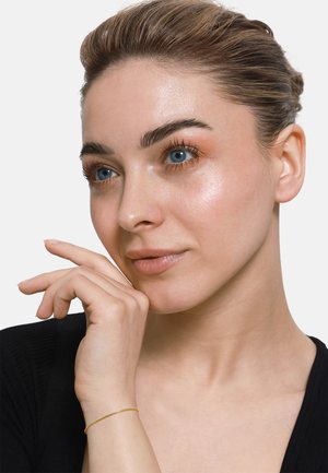 Nahaufnahme einer Person mit heller Haut und blauen Augen, die über glatte Haut, minimales Make-up und ein goldenes Armband am Handgelenk verfügt. Schwarze Kleidung.
