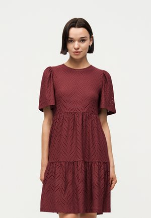 Kvinde med kort mørkt hår iført en vinrød, kortærmet kjole med hulmønster og lagdelte volanter, stående mod en ensfarvet lys baggrund.