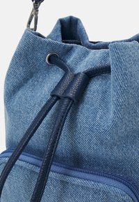 Gros plan d’un sac à cordon en jean bleu avec des cordons en cuir bleu marine et une poche zippée à l’avant.