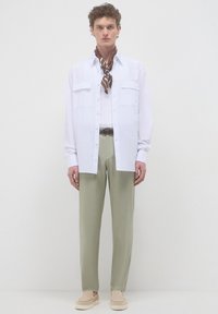 Ein junger Mann steht da, trägt ein weißes offen gehaltenes Hemd, einen braunen gemusterten Halsschal, beige Hosen mit geflochtenem Gürtel und hellbeige Slipper.
