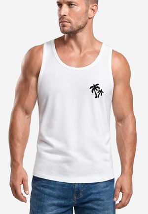 Muscular man wearing a white sleeveless tank top with a small black palm tree graphic on the left chest and blue jeans.