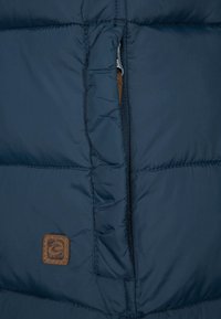 Detalje af mørkeblå dunjakke med syede paneler, en lynlåslomme og en brun lædermærkat med mærkenavn. Glat tekstur.