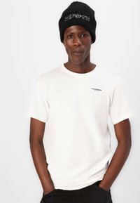 Vit kortärmad t-shirt med rund halsringning och svart logodetalj. Matchad med en svart ribbad basker. Slät bomullstextur.