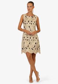 Kantjurk met bloemen uitsparingen in beige en zwart, mouwloos ontwerp, scalloped zoom, gecombineerd met doorzichtige hoge hak sandalen.