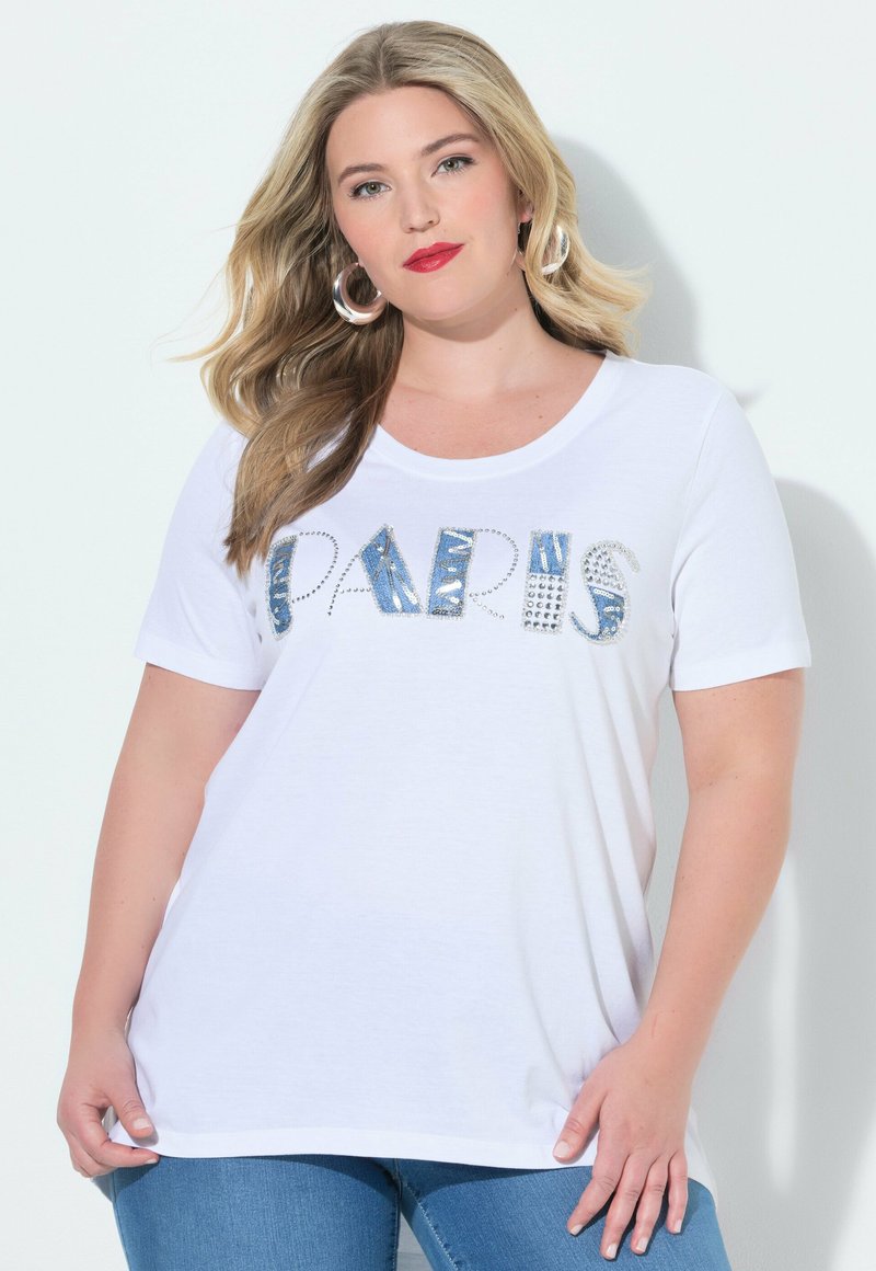 Mujer rubia con lápiz labial rojo, que lleva pendientes de aro, una camiseta blanca con letras "PARIS" tachonadas y vaqueros azules sobre un fondo liso.