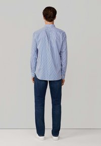 Hombre de pie de espaldas, vistiendo una camisa de rayas verticales azules, jeans oscuros y zapatillas blancas, frente a un fondo liso.