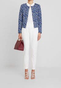 Niebieska kurtka w wzory, biały top, białe spodnie oraz białe sandały na paski. Trzyma bordową torebkę. Gładka faktura i strukturalny design.