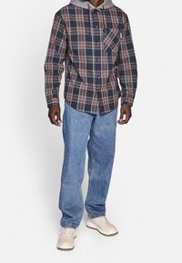 Koszula flanelowa w granatowym kolorze z czerwono-żółtym wzorem w kratę, z szarą kapturem i kieszenią z przodu. Połączona z jasnobłękitnymi dżinsami i białymi butami.