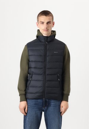 Marinekleurige gewatteerde vest met een rits aan de voorkant, een staande kraag en zijzakken, gedragen over een groene hoodie, gecombineerd met blauwe spijkerbroek.