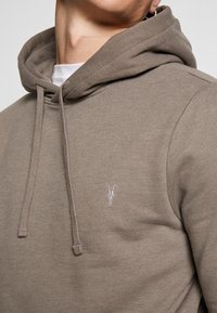 Felpa con cappuccio in taupe con una texture liscia, dotata di una tasca a marsupio frontale e di un piccolo logo teschio ricamato bianco.