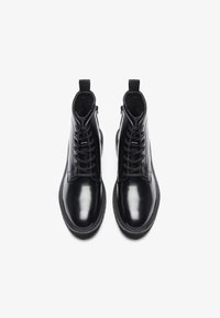 Botas de tobillo de cuero negro con un acabado suave, cordones en la parte delantera, punta circular y tira de agarre en la parte superior. Suela de goma resistente, sin patrones visibles.