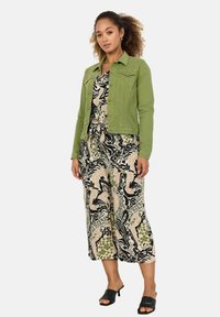 Grön denimjacka över en mönstrad svart och beige jumpsuit med abstrakta mönster, parat med svarta öppen tå-muler.