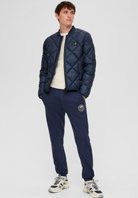 Marineblaue, gesteppte Bomberjacke mit Reißverschluss, darunter ein cremefarbener Pullover, kombiniert mit marineblauen Jogginghosen, die ein Emblem am Oberschenkel haben.