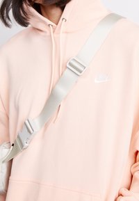 Persikofärgad hoodie med dragsko, vit Nike-logotyp, i kombination med ett brett, ljusgrått justerbart band med spänne i plast.