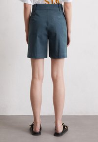 Dark teal cotton shorts with a high waist, side and back pockets, and stitched detailing, ending above the knee. Worn with black flat sandals.