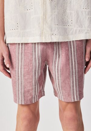 Mann trägt rosa-weiß vertikal gestreifte Shorts und ein weißes Hemd mit floraler Lochstickerei, steht mit entspannt an den Seiten hängenden Händen.