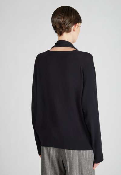 Person mit kurzen Haaren, die ein schwarzes Langarmshirt und graue Nadelstreifen-Hosen trägt, von hinten vor einem schlichten grauen Hintergrund abgebildet.