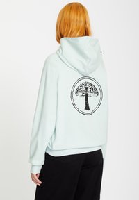 Mintgröna hoodien med en svart trädgrafik i en cirkulär design på baksidan. Har avslappnad passform och ribbade muddar och nederkant.