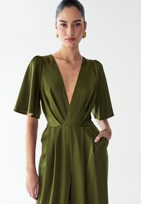 Ženska z nazaj spletenimi lasmi, ki nosi zlate uhane in olivno zeleno obleko z globokim V-izrezom in rokavi do komolcev, z enim rokom v žepu.