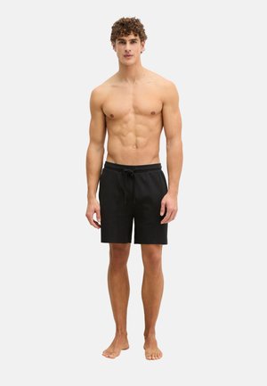 Kale jonge man met krullend haar die zwarte knielange shorts met trekkoord draagt, blootsvoets staand op een effen lichte achtergrond.