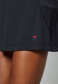 Falda deportiva negra con una textura suave, que presenta un pequeño logo multicolor en el dobladillo. Diseño sencillo con detalles de costuras limpias.