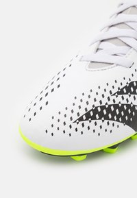 Botín de fútbol sintético blanco con patrones geométricos negros, acentos grises y una suela exterior brillante amarilla. Cuenta con un diseño de cordones.