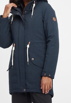 Personne portant une parka bleu foncé à capuche avec des cordons blancs et des poches avant à rabat, ajustant le col d'une main.