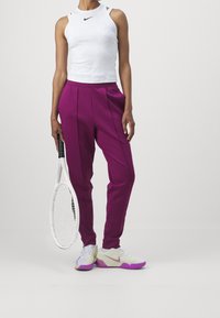 Personne portant un haut blanc sans manches, un pantalon violet et des baskets blanc-violet tenant une raquette de tennis blanche à côté d'un mur blanc.