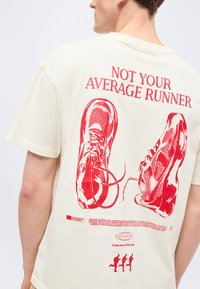 Άνδρας που φοράει μπεζ μπλουζάκι με κόκκινο γραφικό σχέδιο αθλητικών παπουτσιών και το κείμενο "ΟΧΙ Ο ΜΕΣΟΣ ΔΡΟΜΕΑΣ" στην πλάτη.