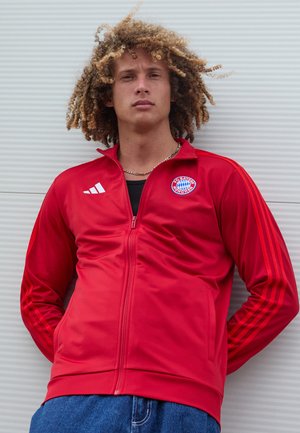 Punane tõmblukuga jakk, millel on kolm valget triipu varrukatel ja FC Bayern Müncheni embleem vasakul rinnal, millel on sile tekstuur.