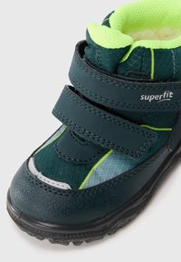 Sötétzöld és neon sárga gyermek téli csizma két tépőzáras pánttal és texturált tapadású talppal, az oldalán "superfit" márkajelzéssel.