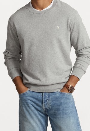 Mand iført lysegrå crewneck-sweatshirt, blå jeans, brunt læderrem-ur, med hænderne i lommerne mod hvid baggrund.