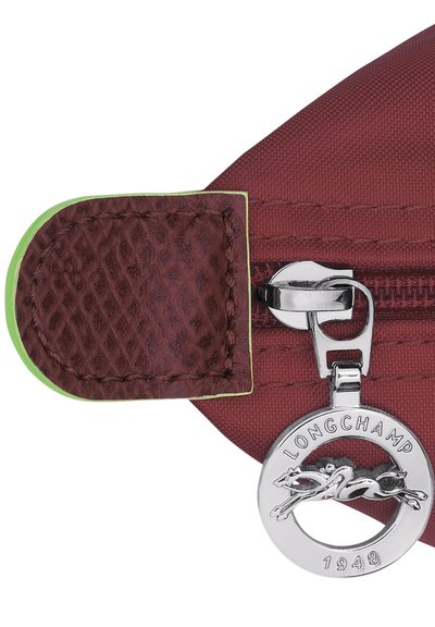 Dark red bag corner with textured leather patch and green trim; silver zipper pull features a round charm embossed with a horse logo and "Longchamp 1948."