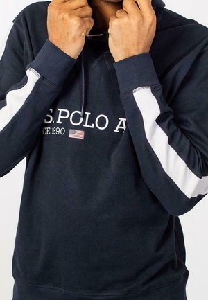 Osoba ubrana w granatową bluzę z kapturem U.S. Polo Assn. z białymi paskami na rękawach, trzymająca sznurki kaptura przy szyi.