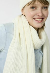 Écharpe en tricot blanche drapée autour du cou, faite de fil doux. Le tissu a une texture de trame lâche, assortie à un bonnet.