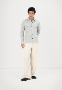 Chemise boutonnée à manches longues grise avec deux poches poitrine, boutons noirs, associée à un pantalon large crème et des chaussures noires. Design simple.