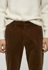 Pantalon en velours côtelé marron avec des côtes verticales, des poches avant et une fermeture à bouton unique. Porté avec un pull crème texturé.