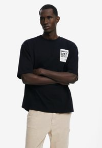 Black t-shirt featuring a rectangular white pocket design with text. Short sleeves and a relaxed fit, made of smooth cotton fabric.