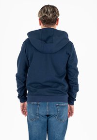 Felpa blu navy con un grande cappuccio, polsini e orlo a costine e una texture liscia. Indossata con jeans di denim azzurri.