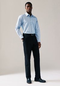 Ljusblå skjorta med ett litet upprepat mönster, button-down-krage, långa ärmar, kombinerad med svarta byxor och blanka svarta skor.