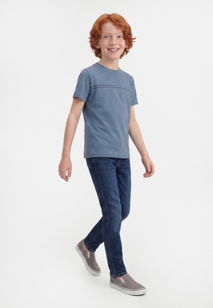 Ragazzo dai capelli rossi che sorride, cammina, indossa una maglietta blu, jeans scuri e scarpe slip-on grigie su uno sfondo bianco uniforme.