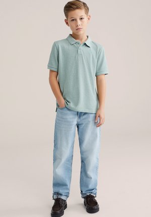 Polo de manga corta de color verde claro, combinado con jeans azul claro y zapatos oscuros. La camiseta presenta un pequeño logo en el pecho.