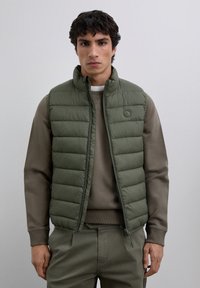 Young man wearing olive green quilted sleeveless vest over brown sweater and matching pants, standing against plain light background.