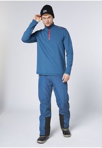 Mann trägt eine schwarze Mütze, ein blaues Langarm-Oberteil mit Reißverschluss, blaue Hose mit schwarzen Aufnähern und schwarze Gummistiefel, steht vor weißem Hintergrund.