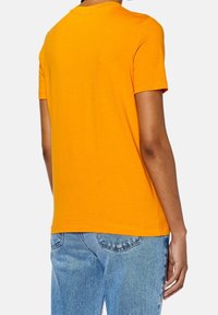 Pomarańczowa bawełniana koszulka z krótkim rękawem i okrągłym dekoltem. Tył jest gładki, bez widocznych wzorów czy akcentów. Noszona z niebieskimi dżinsami.