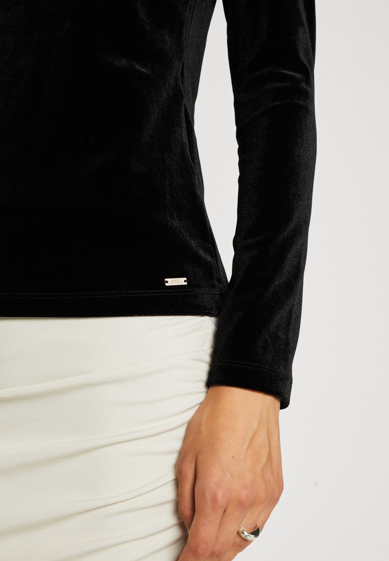 Schwarzes langärmliges Oberteil aus weichem Samtstoff, mit einer figurschmeichelnden Passform und einem kleinen silbernen Etikett am Saum. Kombiniert mit einem weißen Rock.