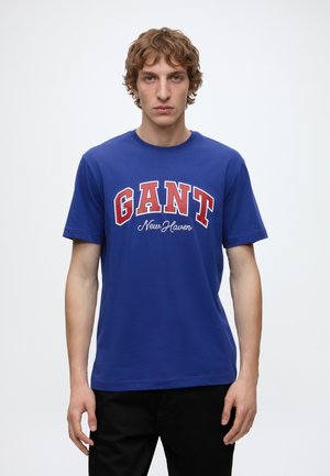 Jeune homme aux cheveux châtain clair bouclés portant un t-shirt bleu avec un logo "GANT New Haven" rouge et blanc, debout devant un fond blanc uni.