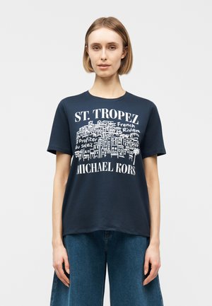 Frau trägt ein marineblaues T-Shirt mit weißem St. Tropez- und Michael Kors-Schriftzug sowie einer Stadtbild-Zeichnung, kombiniert mit blauen Jeans.