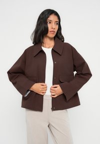 Καφέ, δομημένο σακάκι με γιακά, δύο μπροστινές τσέπες και κλείσιμο με φερμουάρ. Φορεμένο πάνω από λευκή μπλούζα και μπεζ παντελόνι. Ομαλή υφή υφάσματος.